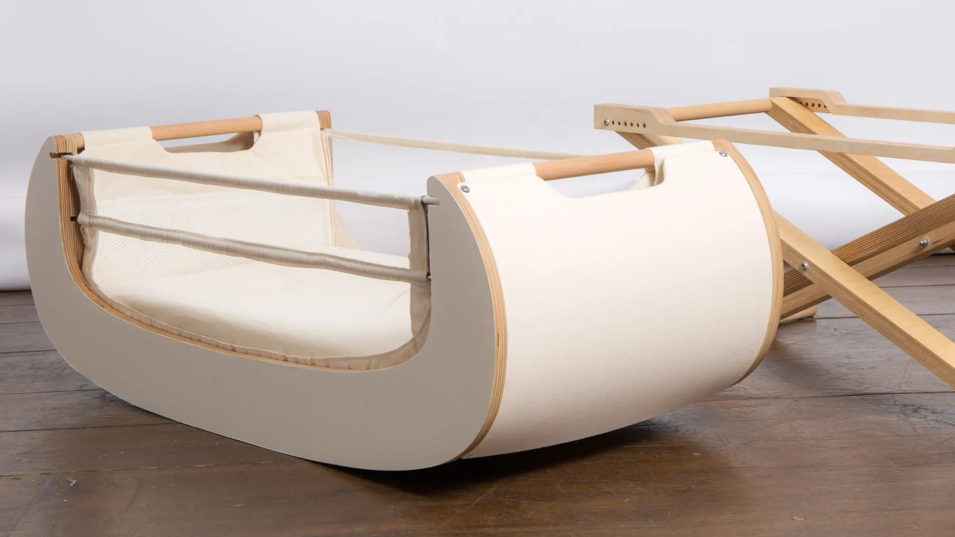 The Alora crib body lifted off its stand on a wooden floor, showing the curved rocking base, canvas sides and mesh panel, with the folded stand behind it.