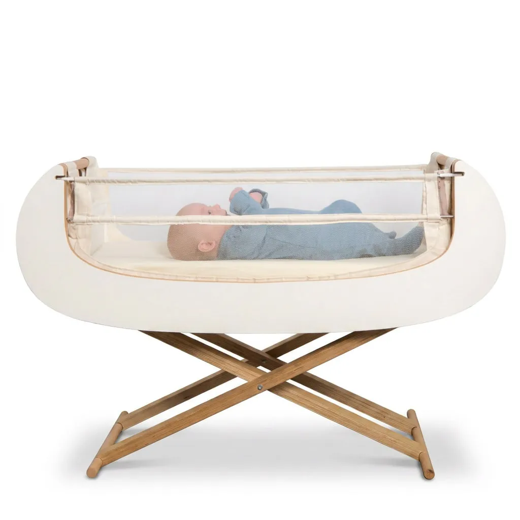 The Alora bedside crib on its folding wooden scissor stand, photographed straight on, with a baby in a blue sleepsuit lying inside behind the mesh side.