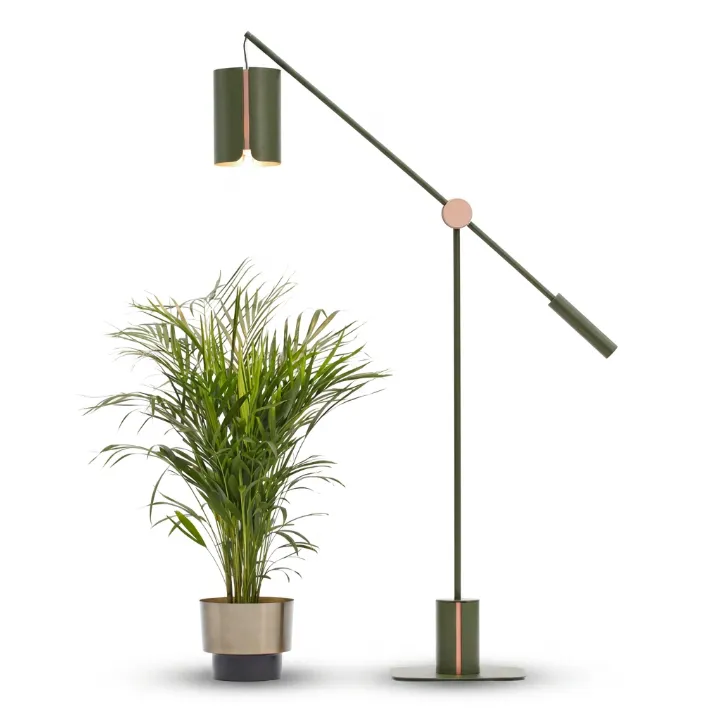 The Anthos grow lamp, an olive green counterbalanced floor lamp with a copper pivot and a weighted disc base, its cylindrical shade angled over a potted palm.