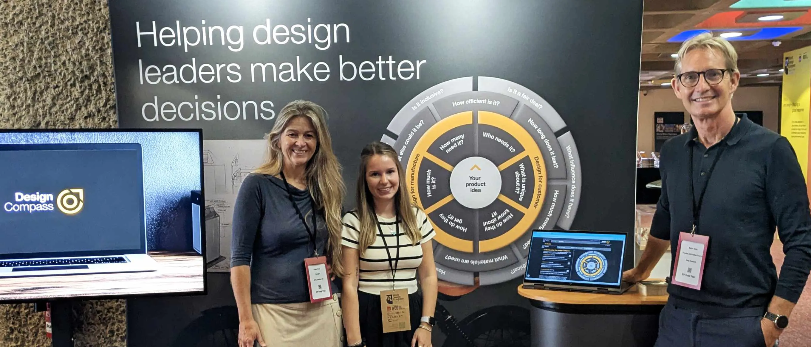 Three of the Bang Creations team standing at their Design Compass exhibition stand, which carries the line Helping design leaders make better decisions beside a large printed Design Compass wheel.