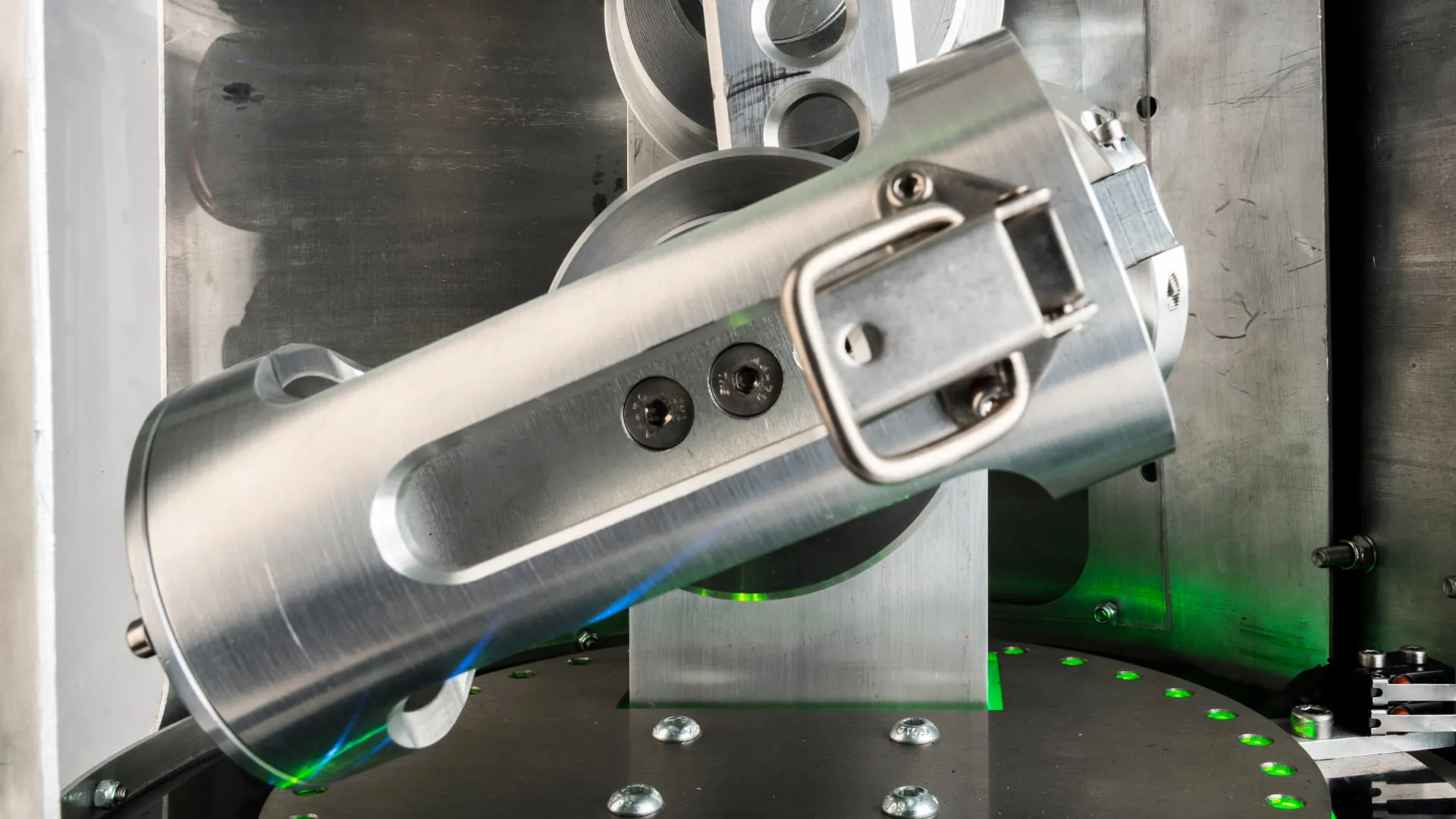 Close up inside the mixer of a machined aluminium cylinder held at an angle by a sprung clamp, above the green lit turntable.