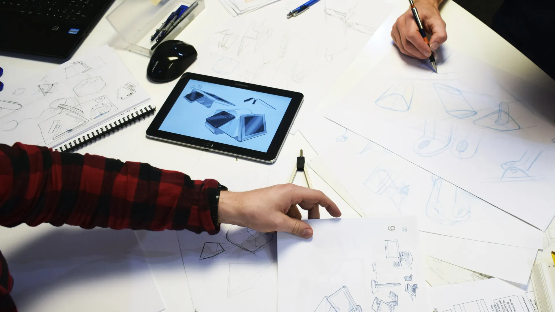 An overhead view of a concept session: two designers sketching across a desk of product drawings, with a tablet showing early blue 3D renders between them