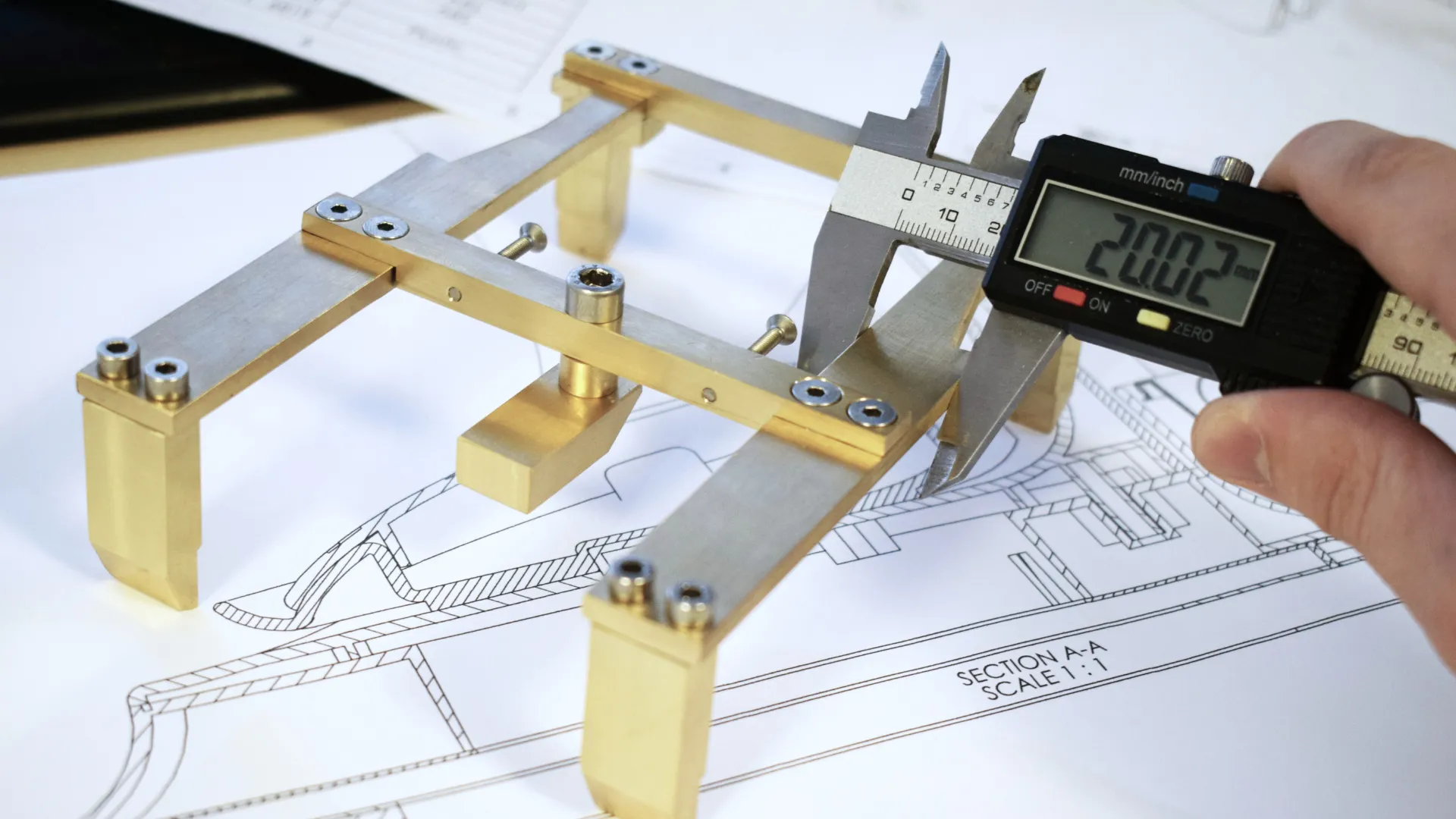 Digital callipers measuring a machined brass fixture on top of its 1:1 engineering section drawing