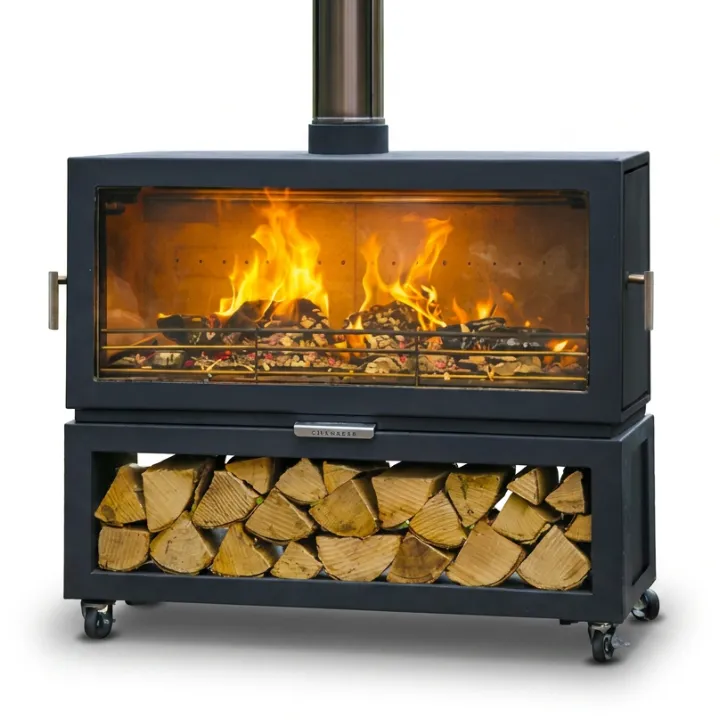 The Chesneys outdoor stove alight, a wide black rectangular firebox with a full width glass front and a flue on top, standing on castors over a log store stacked with cut logs.