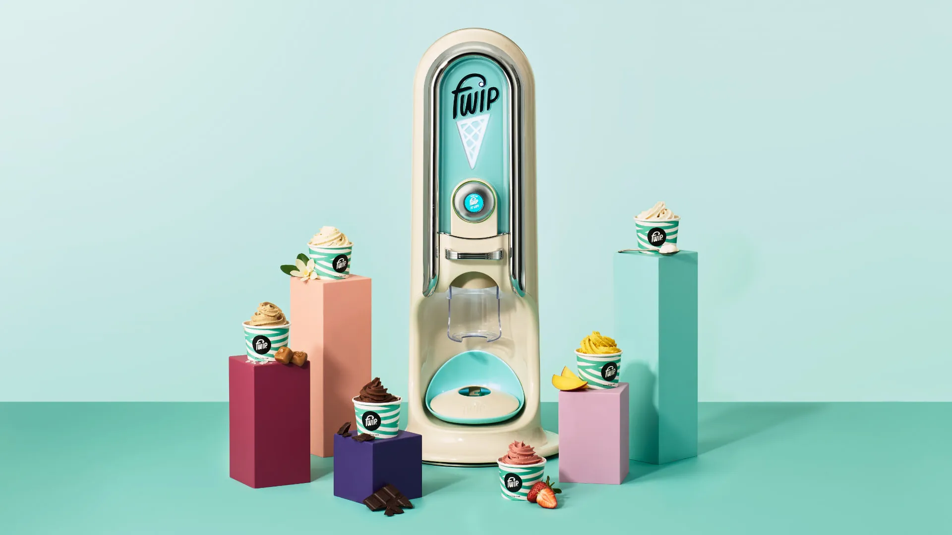 The cream and turquoise machine on a mint studio background, surrounded by striped Fwip tubs of vanilla, caramel, chocolate, strawberry and mango dessert on coloured plinths.