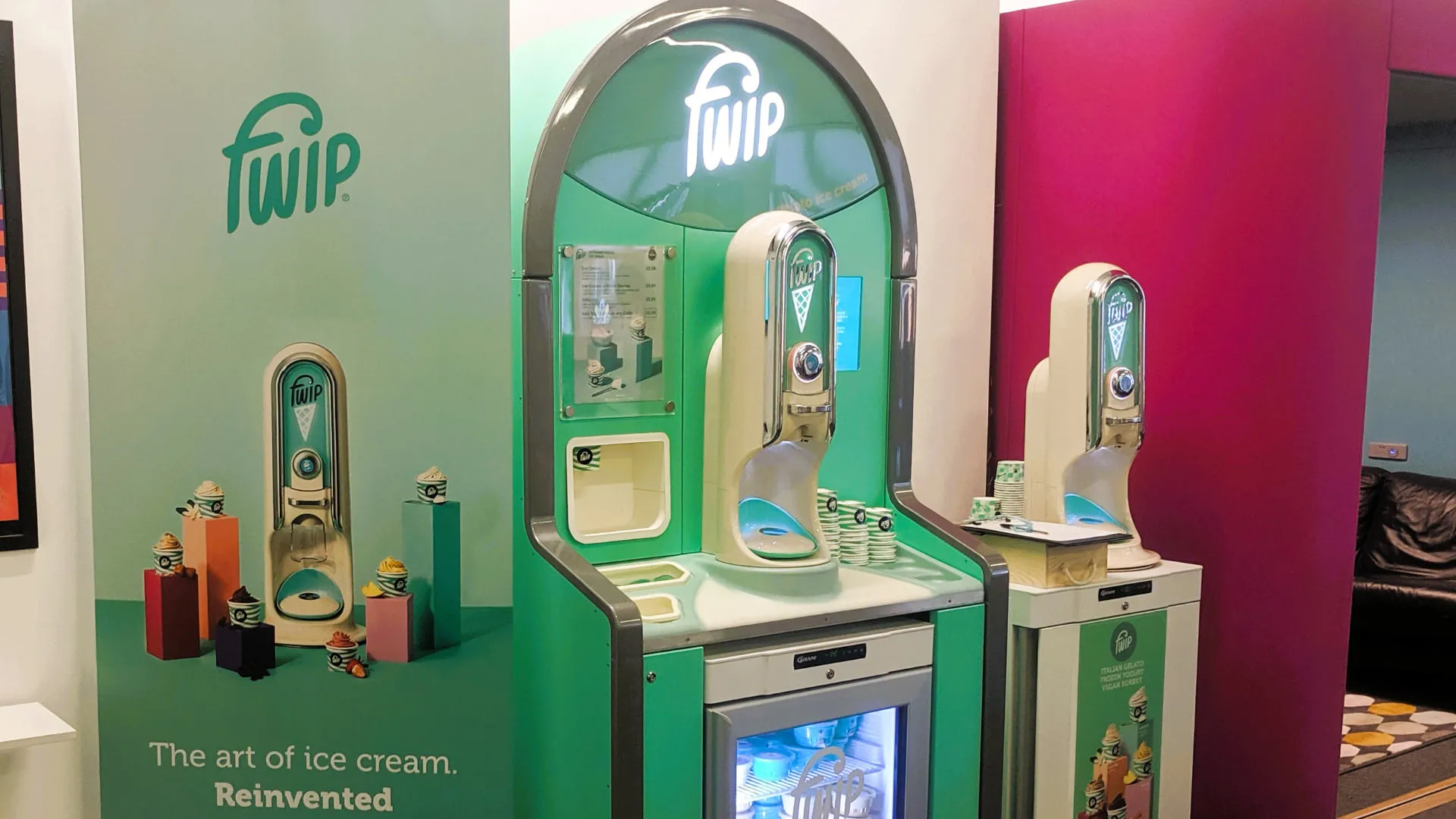 Two machines installed in a retail space, one built into a green branded stand with a chilled pod cabinet below and a menu board, beside a banner reading The art of ice cream. Reinvented.