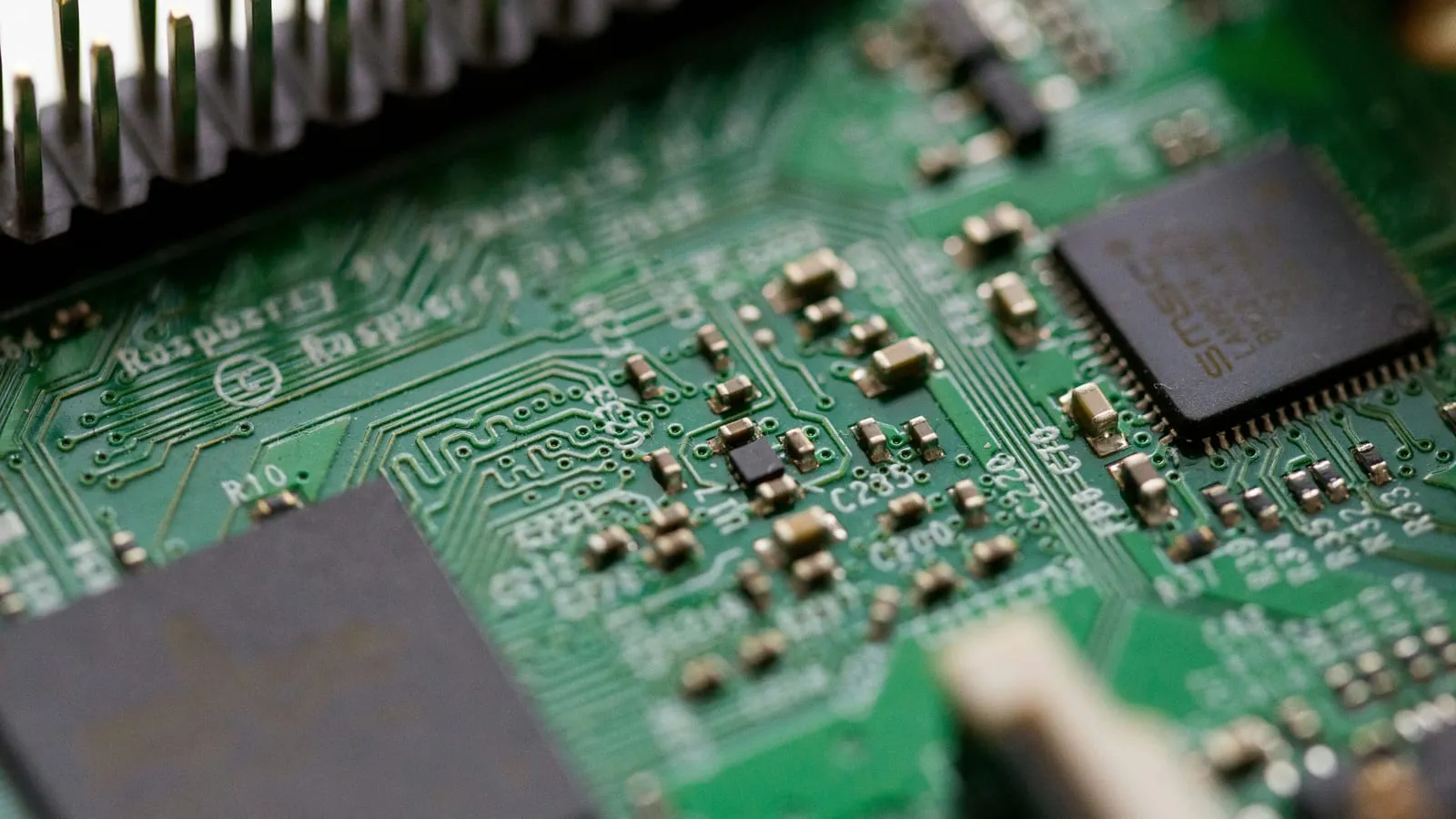 Macro close-up of a populated green circuit board, the Raspberry silkscreen legible beside the pin header, with surface-mount components and a large chip in shallow focus.