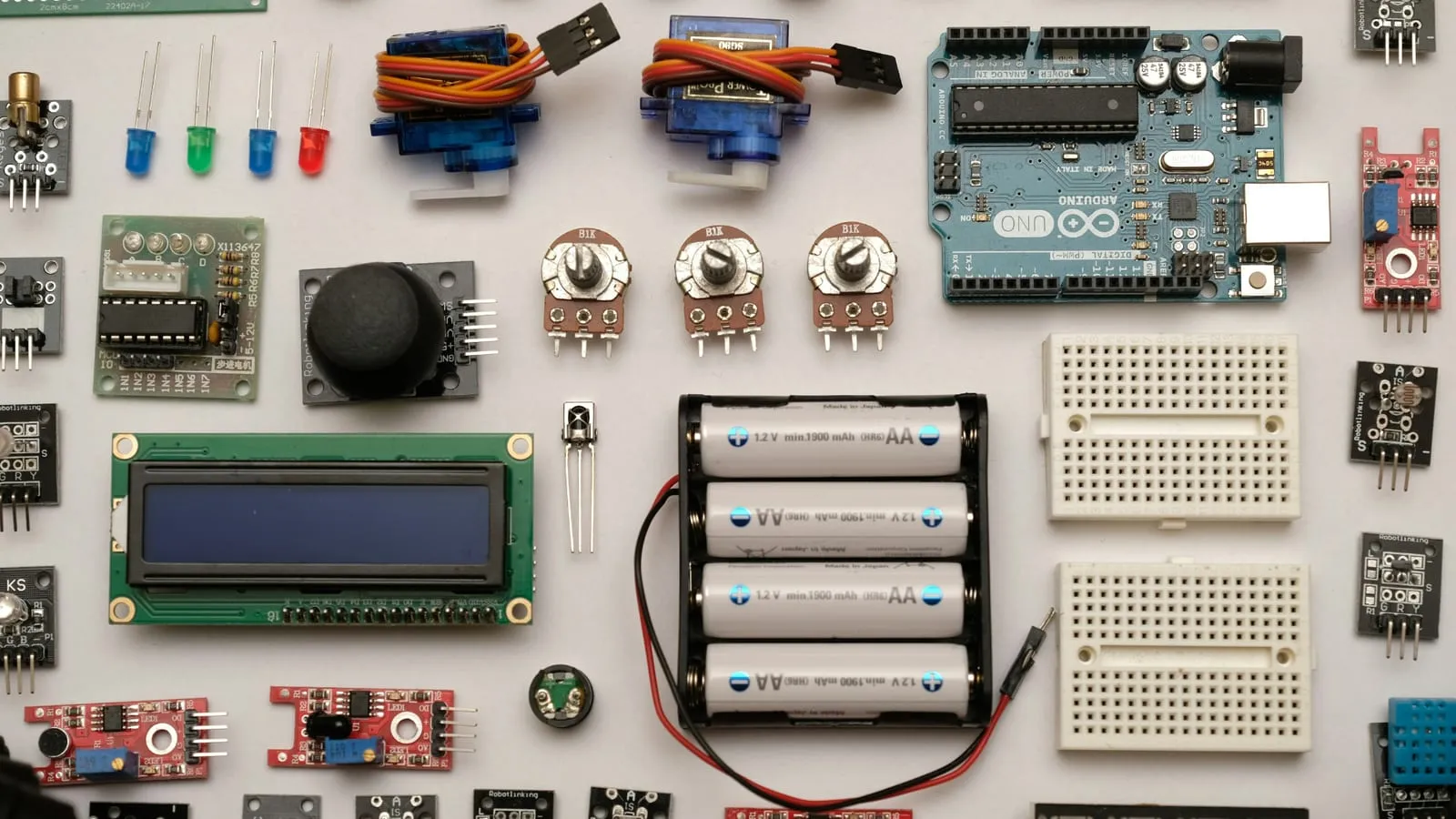 A flat lay of off-the-shelf prototyping electronics arranged in rows: an Arduino Uno, two servos, coloured LEDs, potentiometers, a joystick, an LCD module, breadboards, a AA battery holder and a range of small sensor boards.