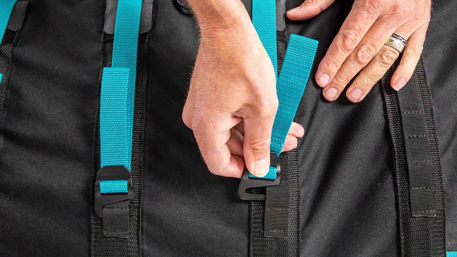 Close-up of two hands feeding a turquoise webbing strap through a black hook buckle on the bag's black technical fabric, tensioning one rib of the internal protection system.
