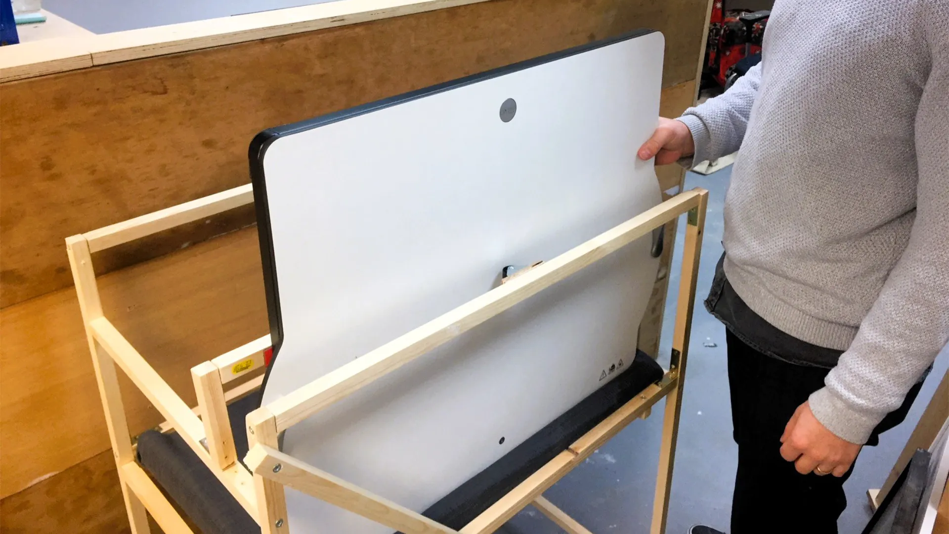 A hand lifting the white Opløft desktop out of its black tube-frame storage stand in a workshop, revealing the yellow warning label and the OPLØFT lift and carry instruction graphics.