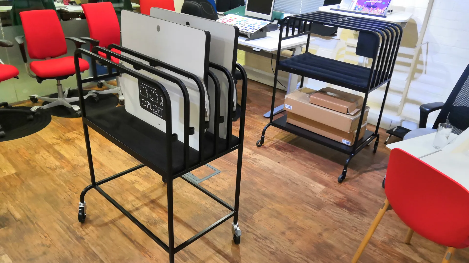 Two loaded Opløft storage stands on castors in an office, one holding a stack of desktops labelled with the OPLØFT lift instructions, the other stacked with cardboard delivery boxes.