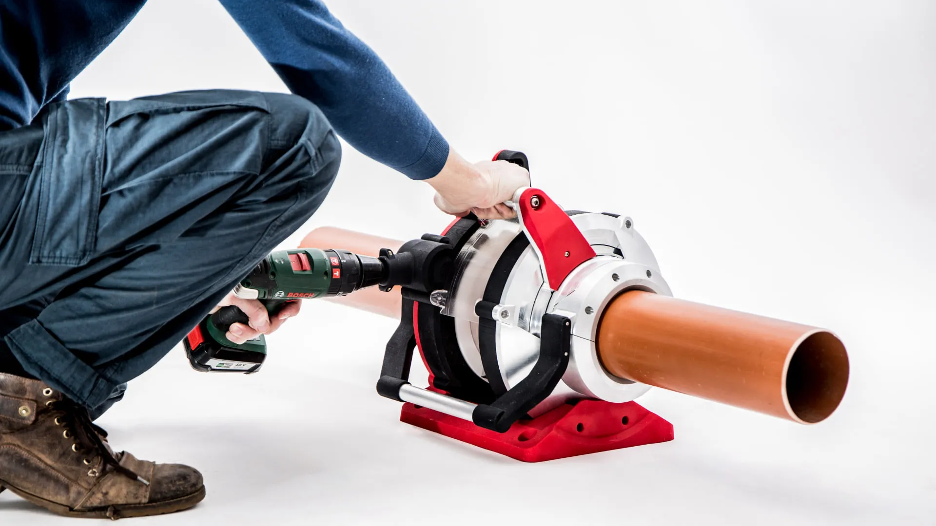 A kneeling worker in overalls driving the tool with a cordless drill, one hand on the tool's handle, as it cuts a brown drainage pipe held in its clamp.