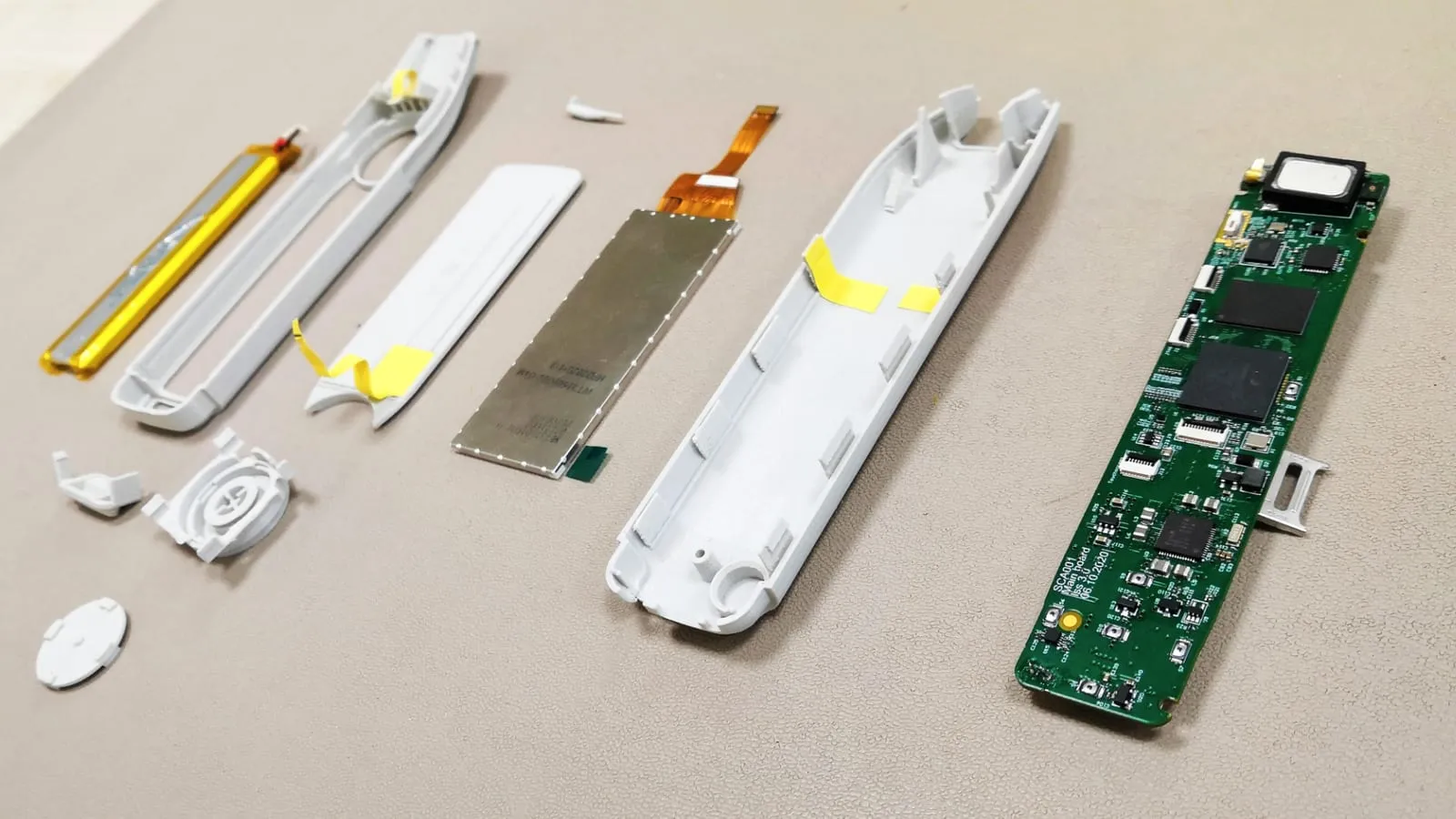 The parts of a slim white handheld device laid out in order on a bench: battery, moulded housing halves, screen, buttons and the populated green circuit board.