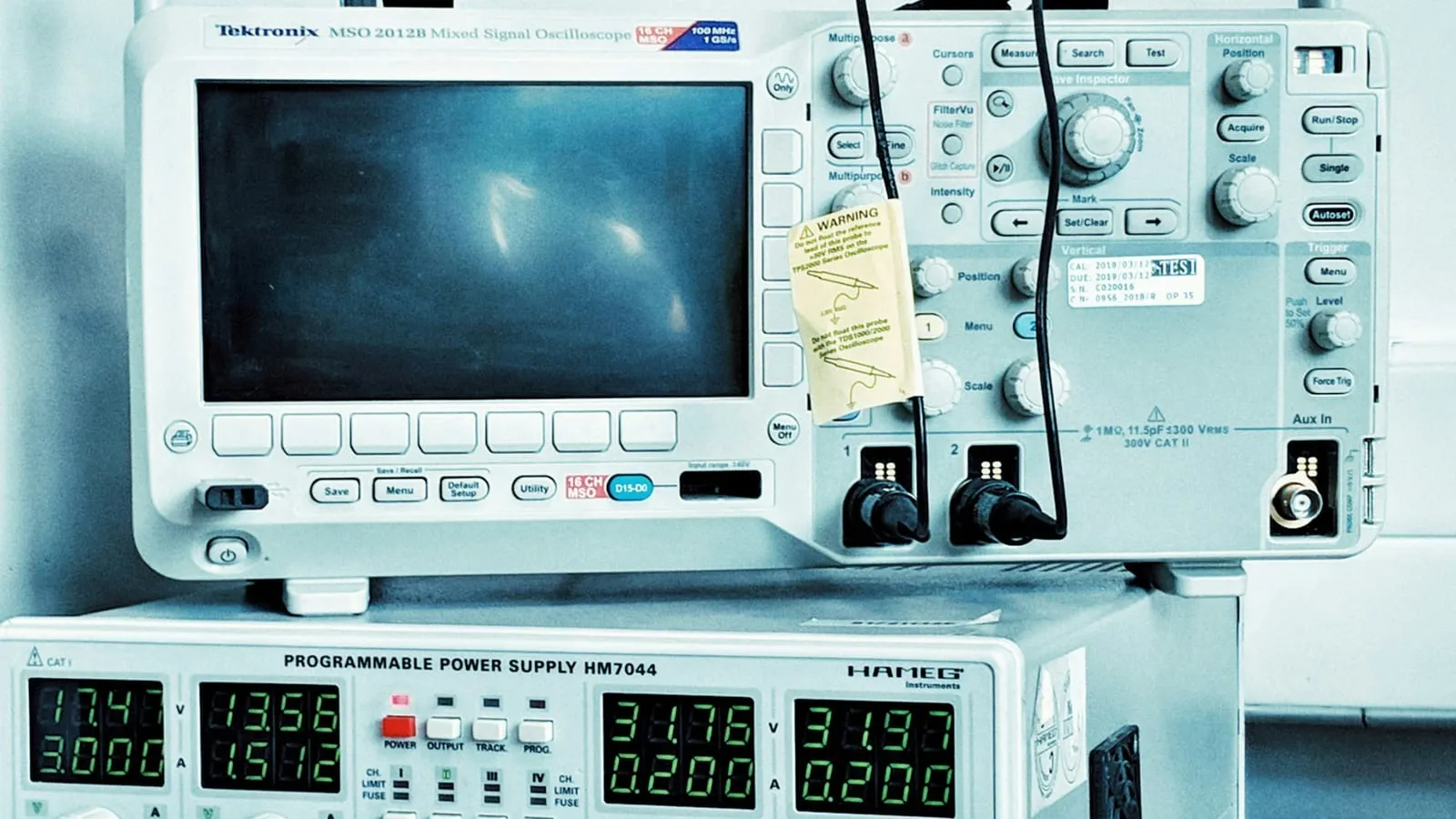 A Tektronix mixed signal oscilloscope carrying a calibration sticker with test and due dates, stacked on a programmable bench power supply showing voltage and current readouts.