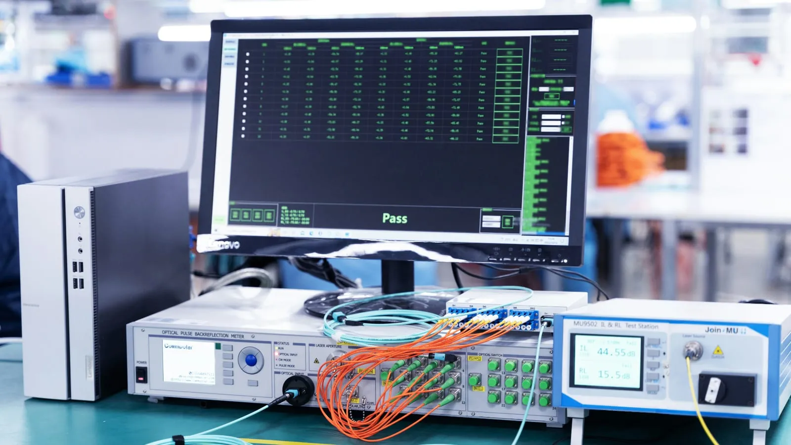 A test house bench with a monitor showing a table of measurement results, every row marked Pass in green, in front of an optical pulse backreflection meter and a test station wired up with orange and blue fibre patch leads.
